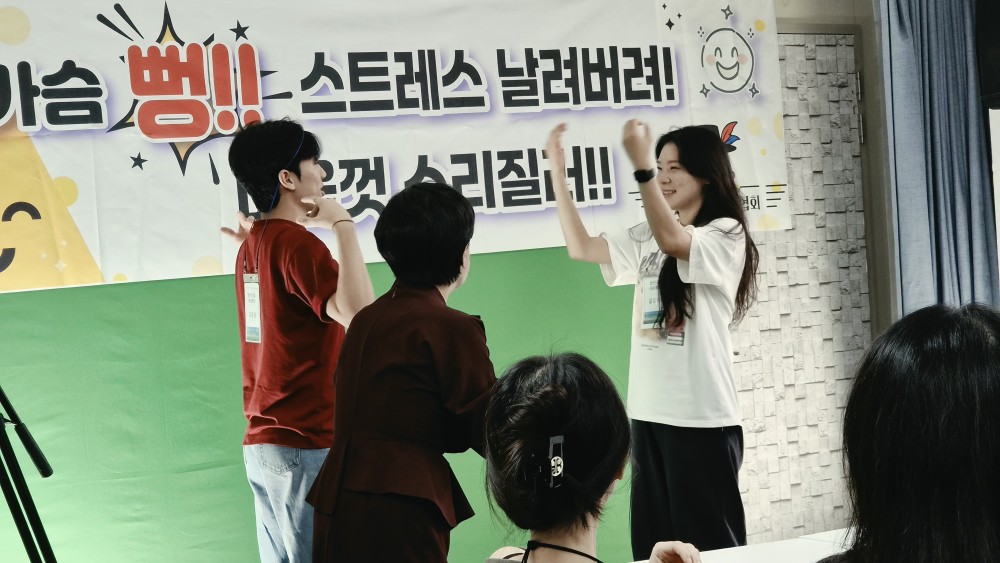 강사가 마이크를 들고 있고 참가자들이 두 손을 들어 동작을 따라 하고 있다