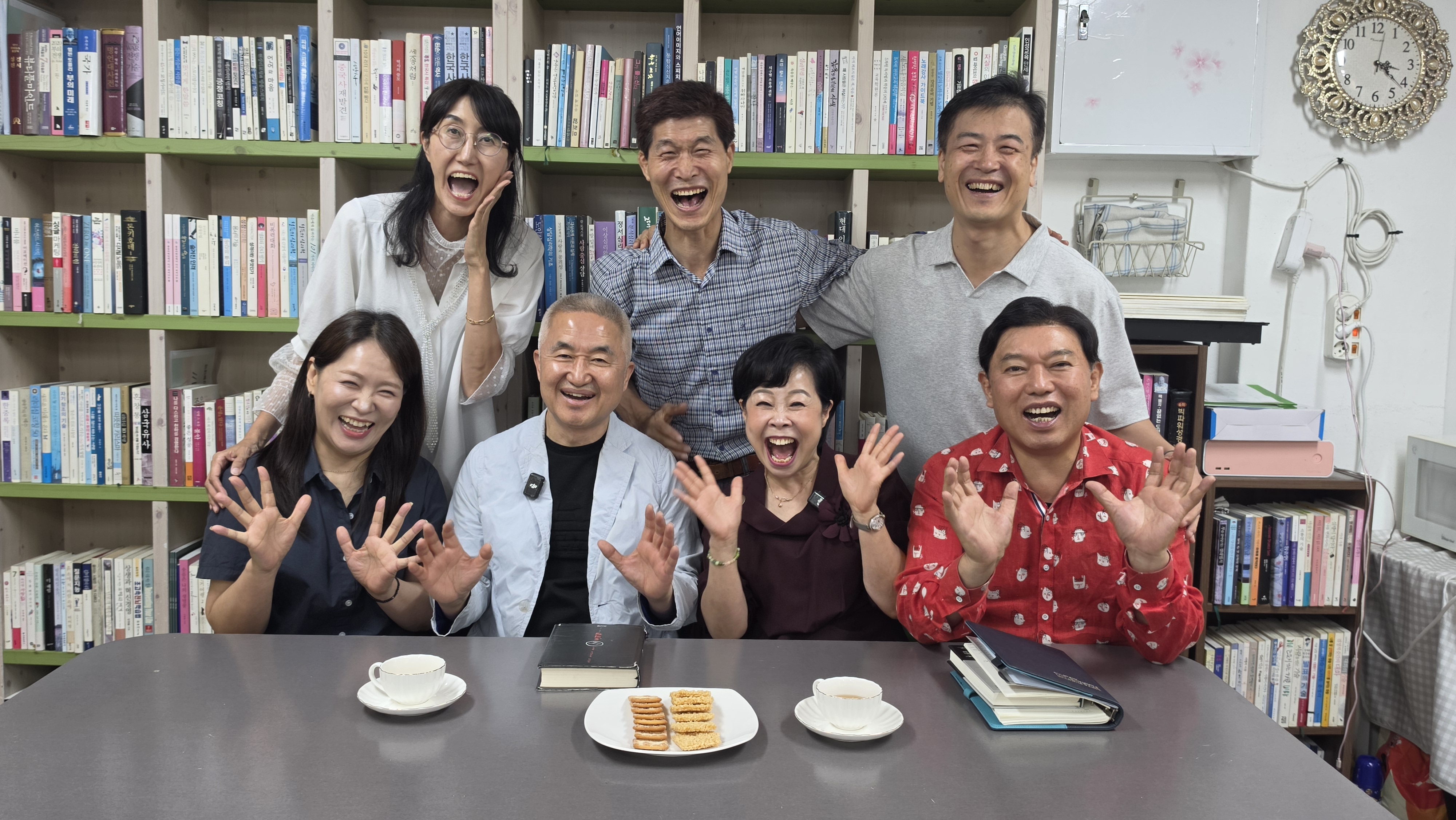 책장 앞에서 참석자 일곱 명이 손을 들고 입을 크게 벌려 웃으며 찍은 단체 기념사진